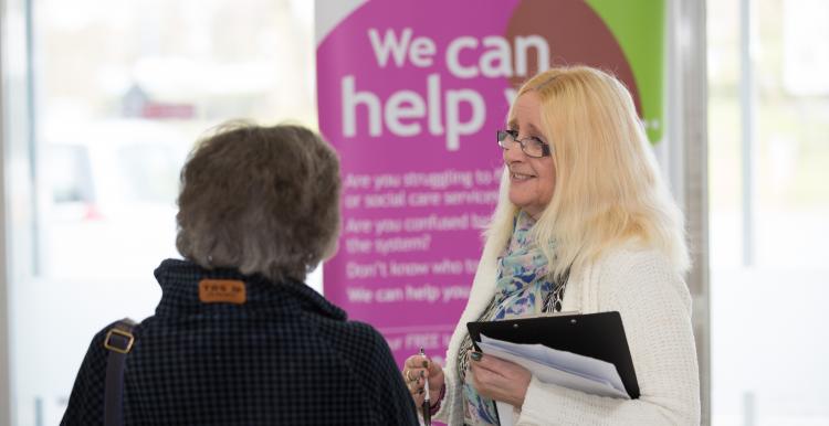 Woman standing in front of a banner that says 'we can help' speaking to a member of the public