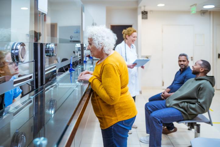 Senior woman talking with receptionist in waiting room in medical clinic. Two men talking with female doctor in the background.