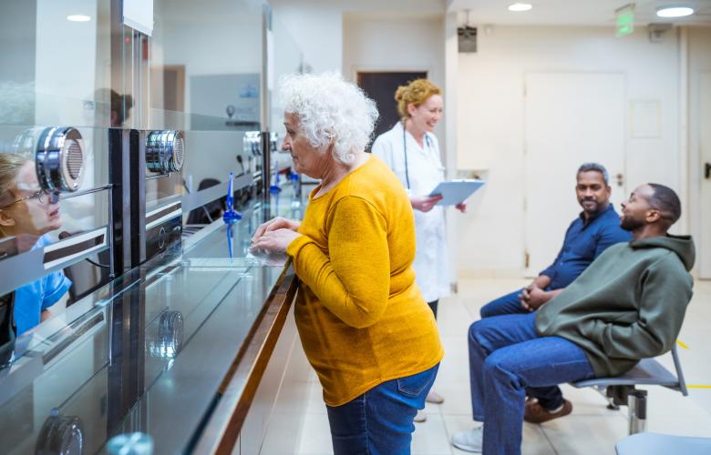 Senior woman talking with receptionist in waiting room in medical clinic. Two men talking with female doctor in the background.